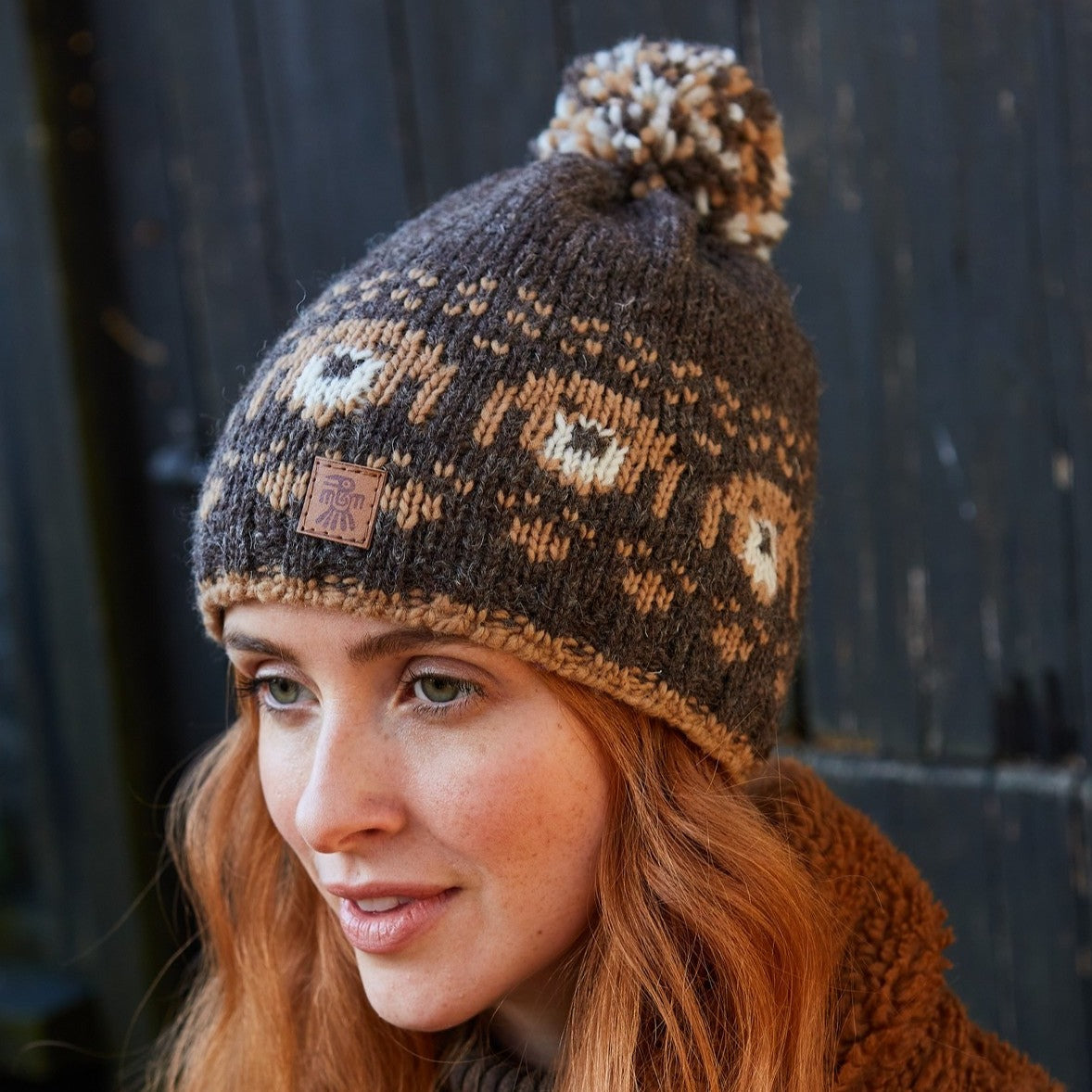 A woman wearing a brown and beige knitted wool bobble beanie hat with a dog pattern and a pom-pom on top.