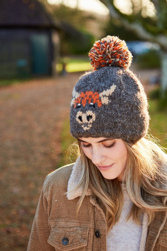 A woman wearing a hand knitted brown wool beanie with a pattern of a Highland cow, lined with fleece around the forehead.