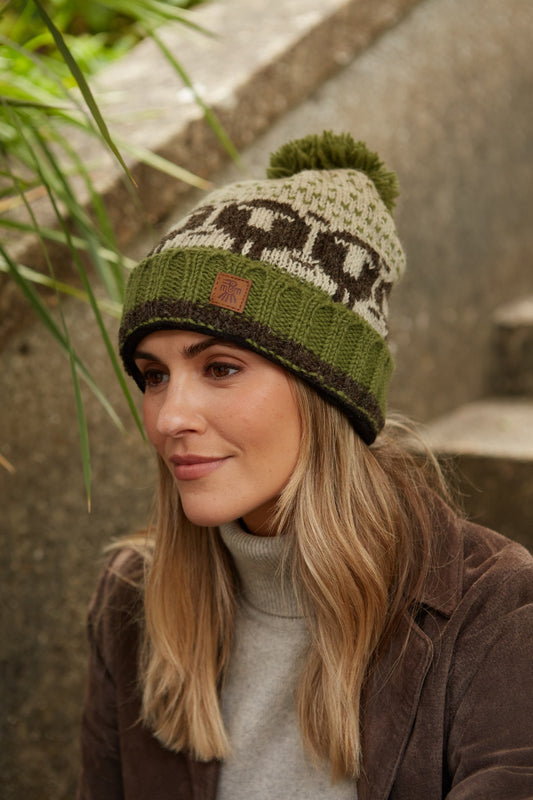 A person wearing a green, brown, and beige striped beanie with a bobble pattern, matching a handwarmer, sitting outdoors.