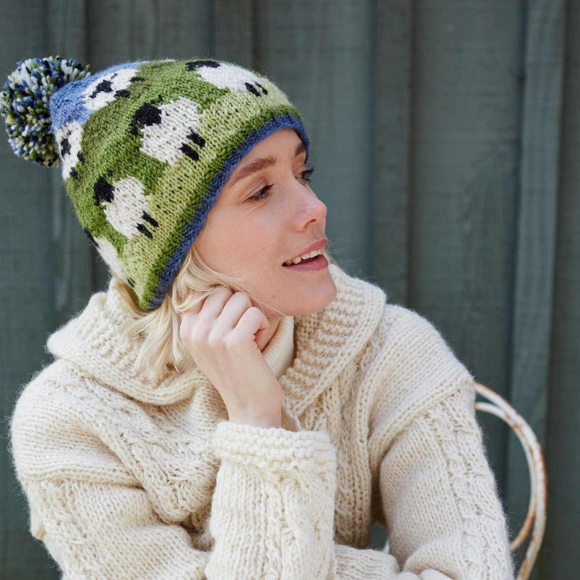 A person wearing a green and white bobble beanie hat with a sheep design and a blue pom-pom.